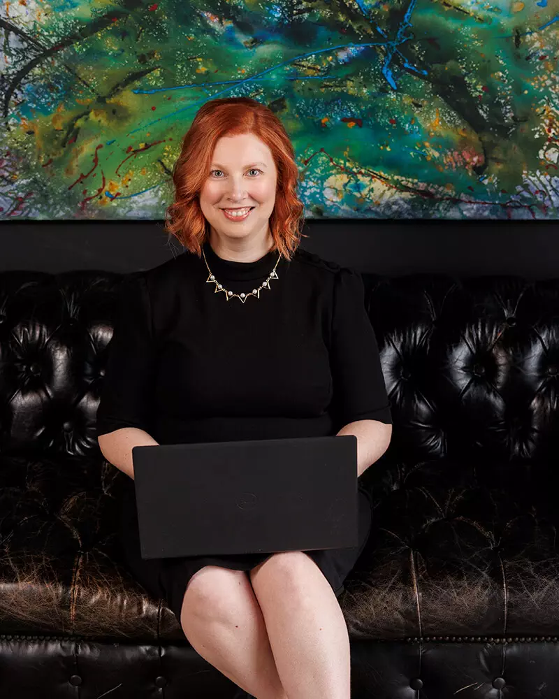 redhead young woman in black dress on leather couch with laptop